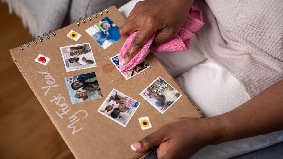 A woman holds a scrapbook photo album with pictures of a little girl on her own or with her parents and a title saying ‘My first year’ on her lap and wiping water droplets off of one of the pictures with a bright pink cloth.