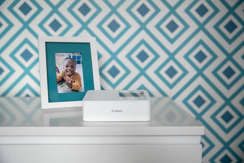 A white SELPHY CP1500 on a white chest of drawers with a picture of a little girl smiling in a white frame standing next to it. A graphic blue wallpaper is in the background.