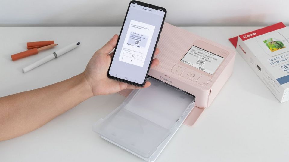 A hand holding a phone scanning a QR code from the pink SELPHY CP1500 printer LCD screen. SELPHY paper packaging is visible in the right corner and a pen, and a pencil are visible in the left corner.